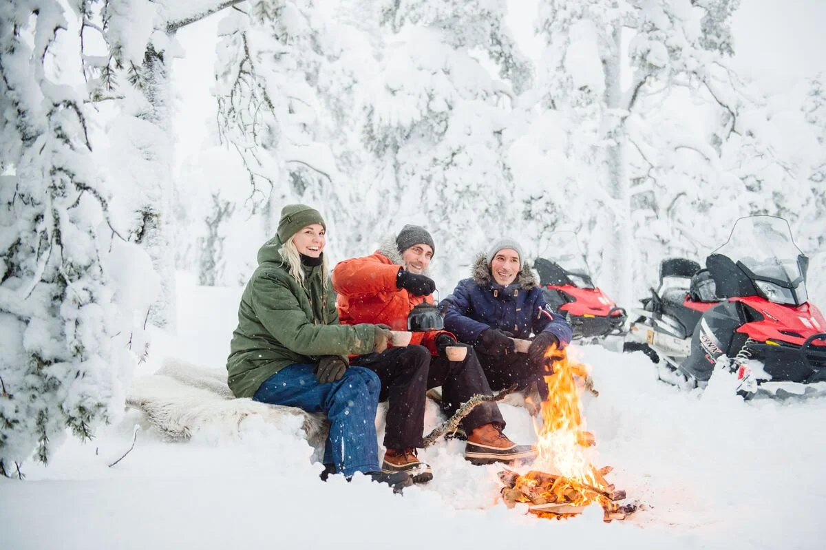 Three colleagues laughing over coffee at an open fire in a snow-covered forest, snowmobiles parked behind them