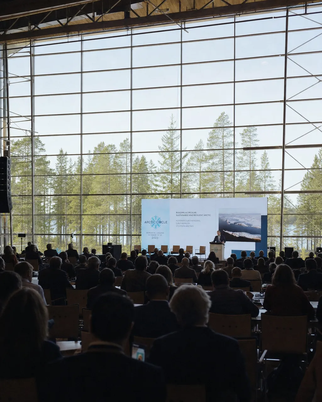 Inside a large glass-domed Arctic auditorium, delegates seen from behind as silhouettes against a backlit professional stage, surrounding pine forest visible through the dome