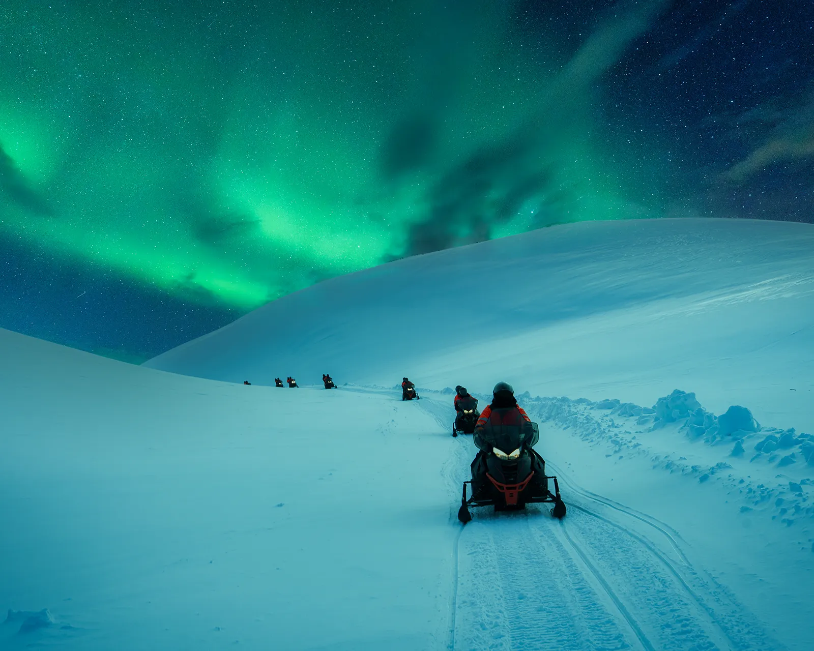 A line of snowmobiles led by a single rider climbs an open snow ridge in Finnish Lapland beneath a bright green aurora and a star-filled sky