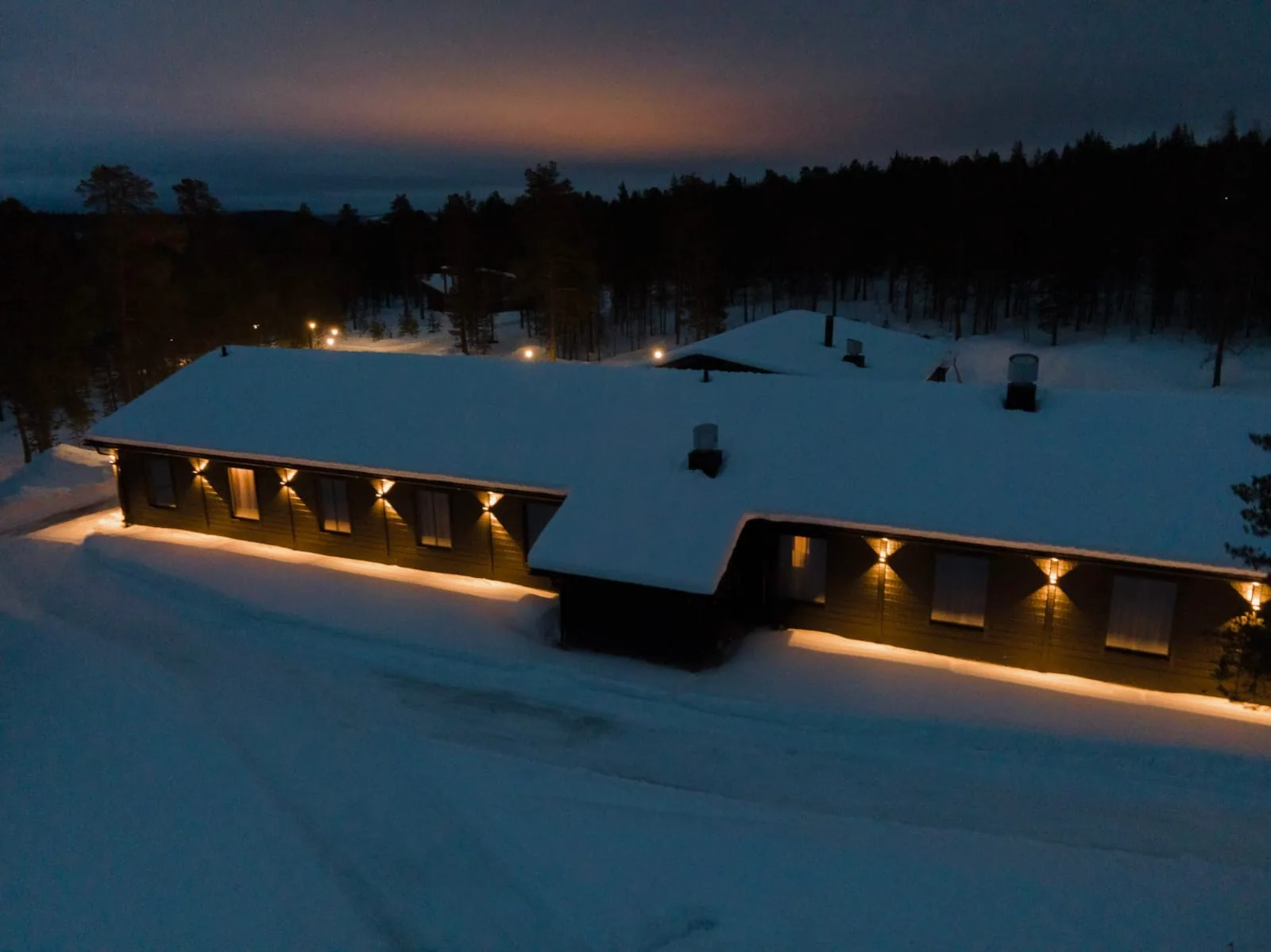 Hotel Kuura from above at dusk: snow lying on the roof, warm lights under the eaves running the length of both wings, forest and a dusky orange sky behind.