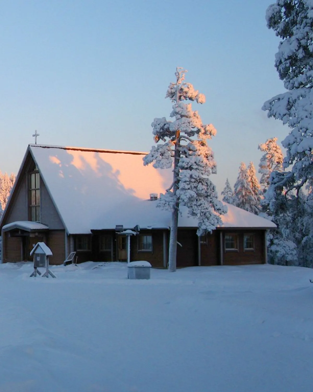 Pyhän Paavalin Kappeli at Saariselkä — a wooden fell chapel under thick snow at golden hour, snow-laden pine in centre frame